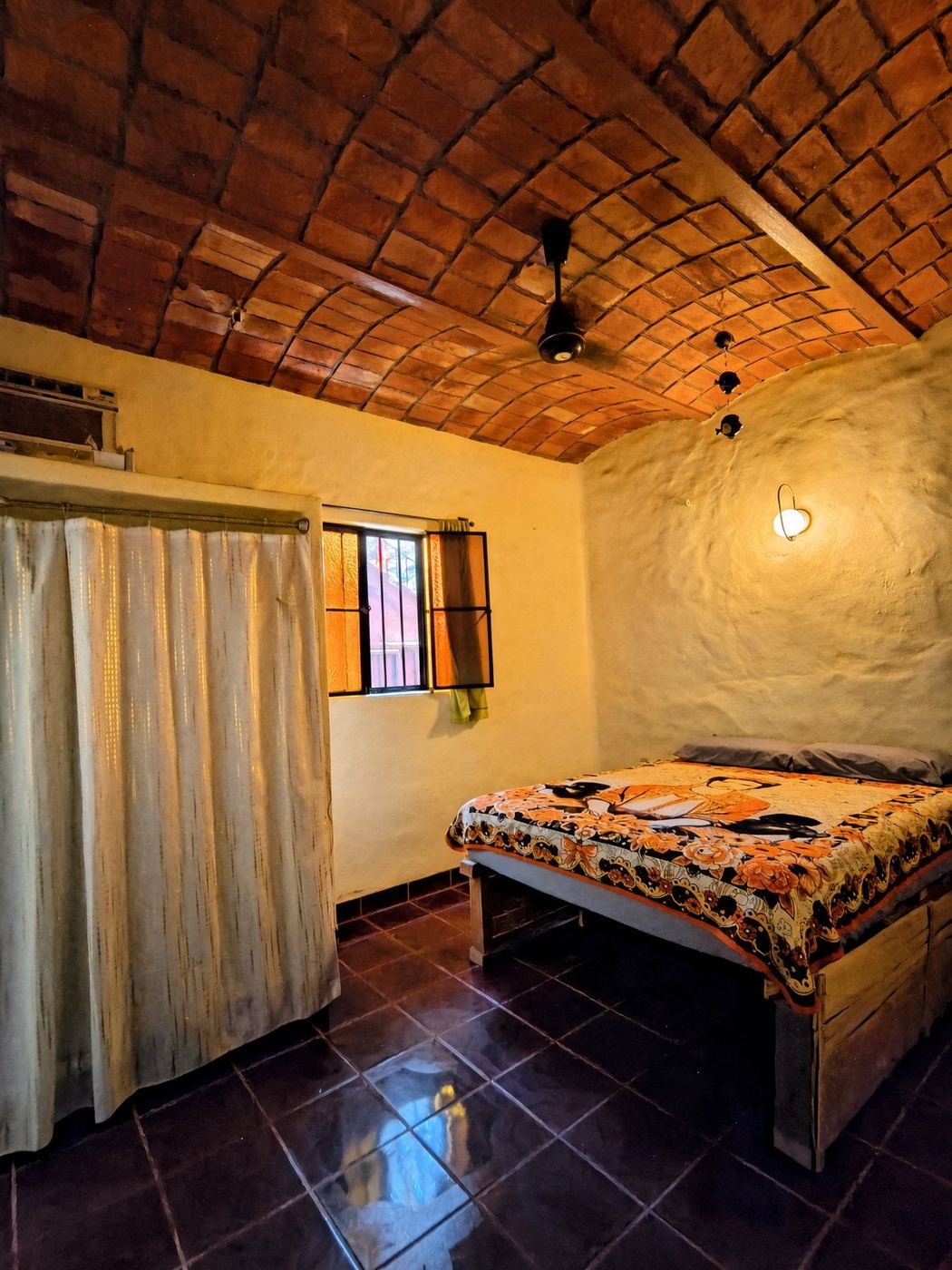 A bedroom at La Casa Grande, floral quilt, sheer curtain, and a warm brick ceiling